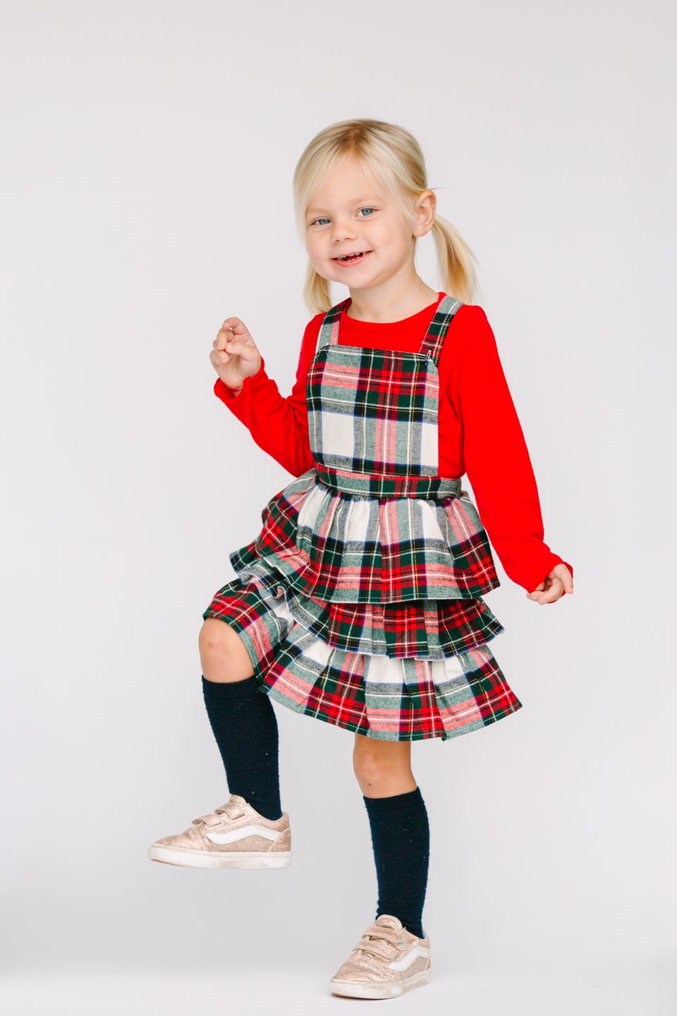 Little girl in a red top and plaid jumper kicking up one foot in the studio