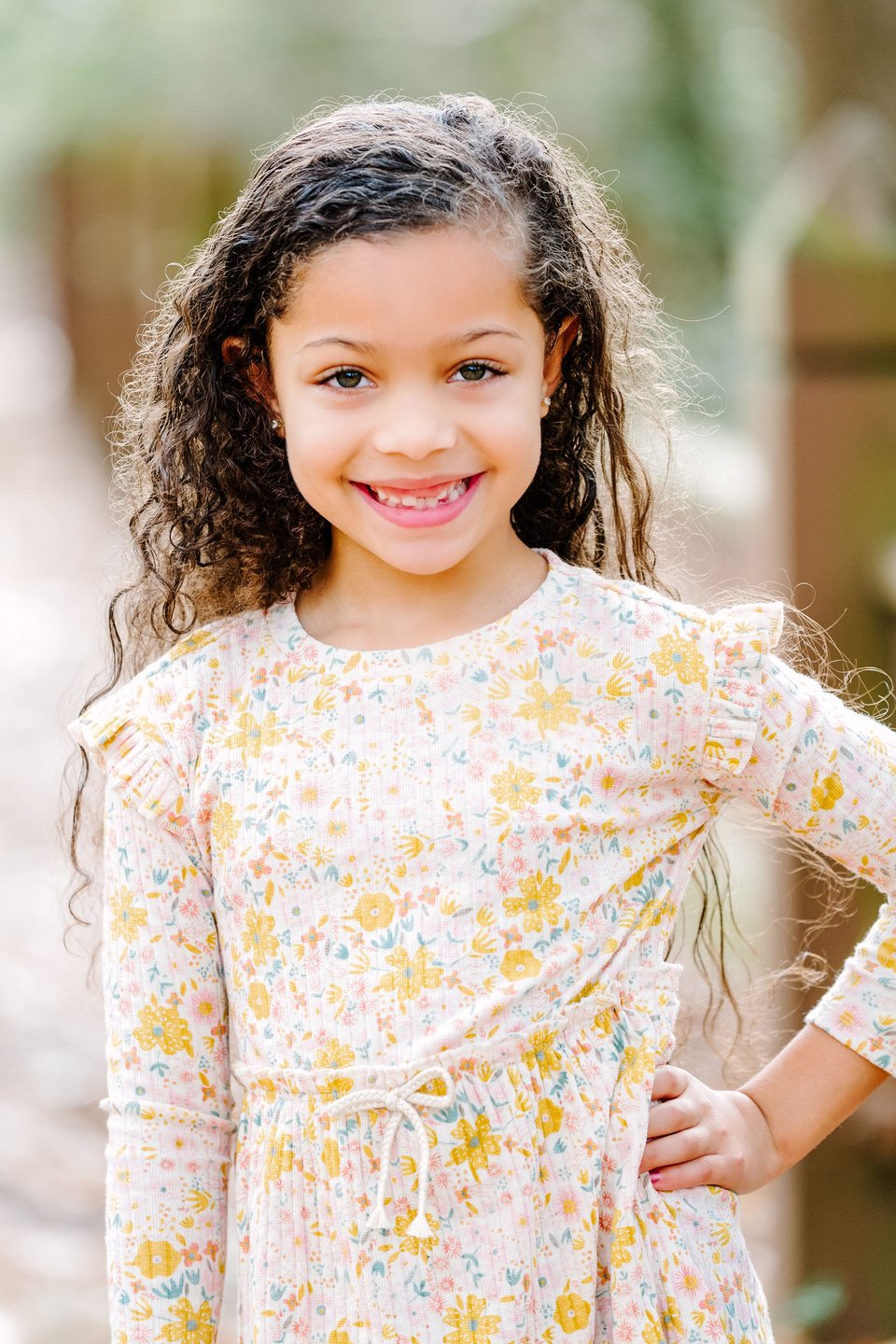 Child in a yellow floral dress with a hand on her hip on a leafy path