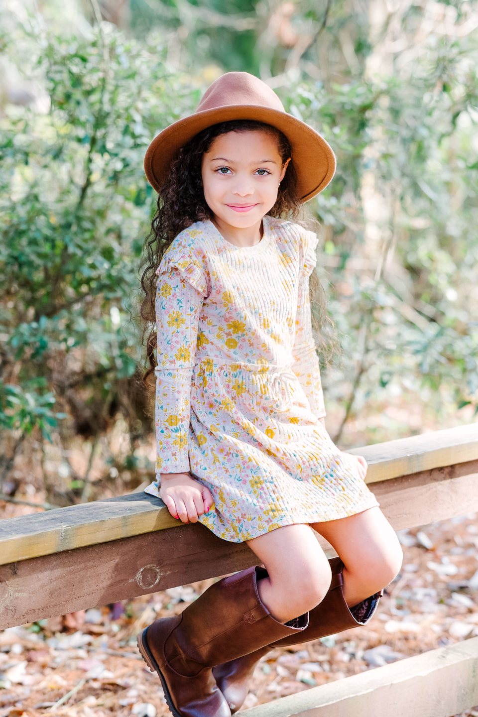 Child in a floral dress and brown hat sitting on a wooden railing