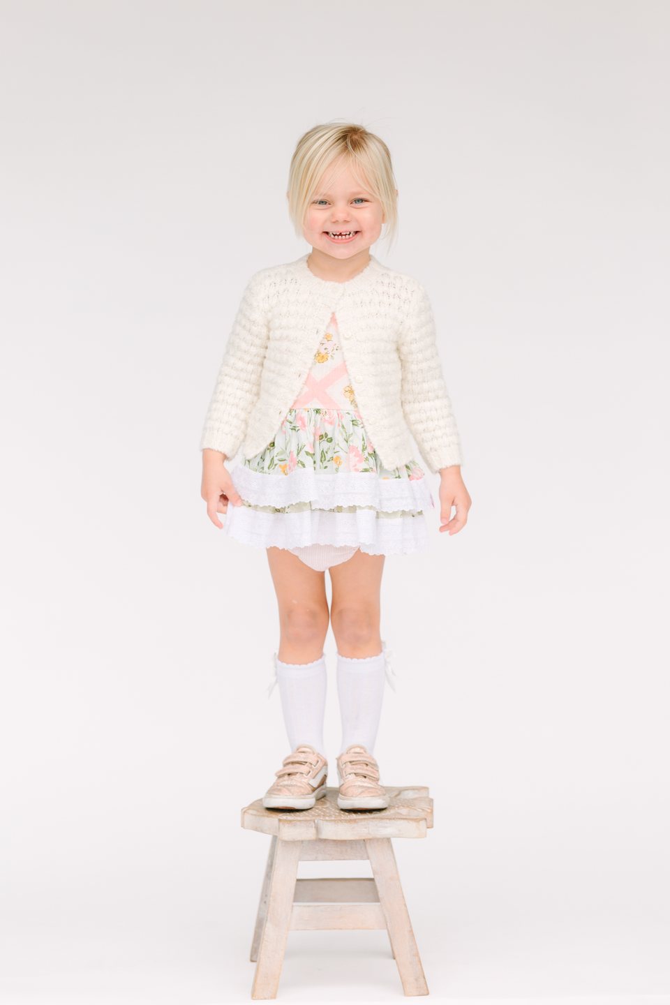 Toddler in a cream cardigan standing on a small wooden stool in the studio