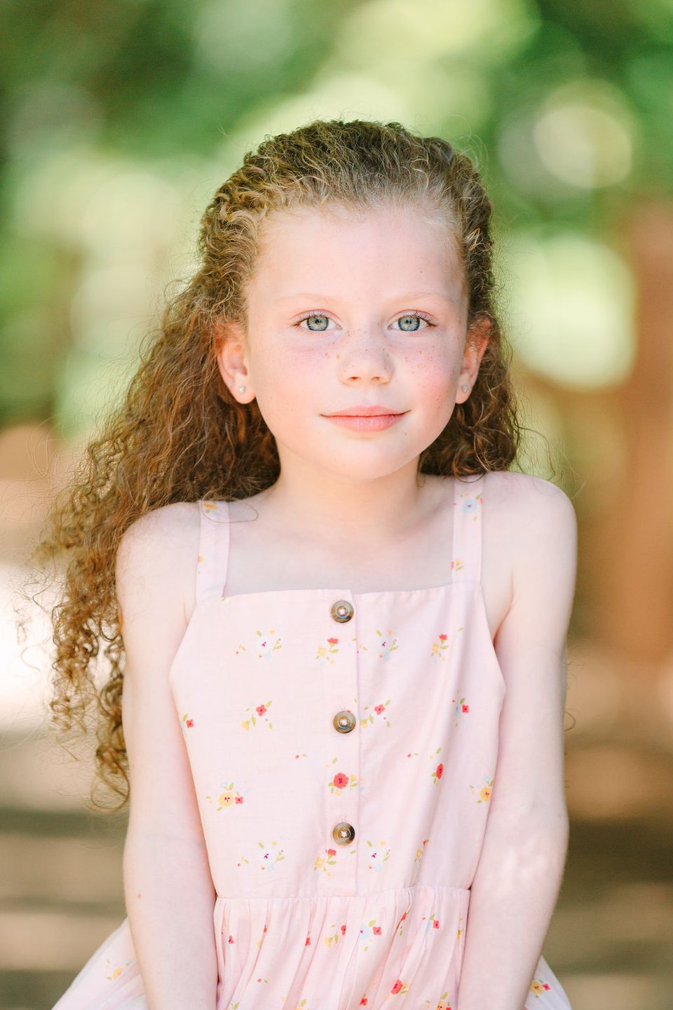Girl with curly red hair in a pink sundress smiling softly among green trees