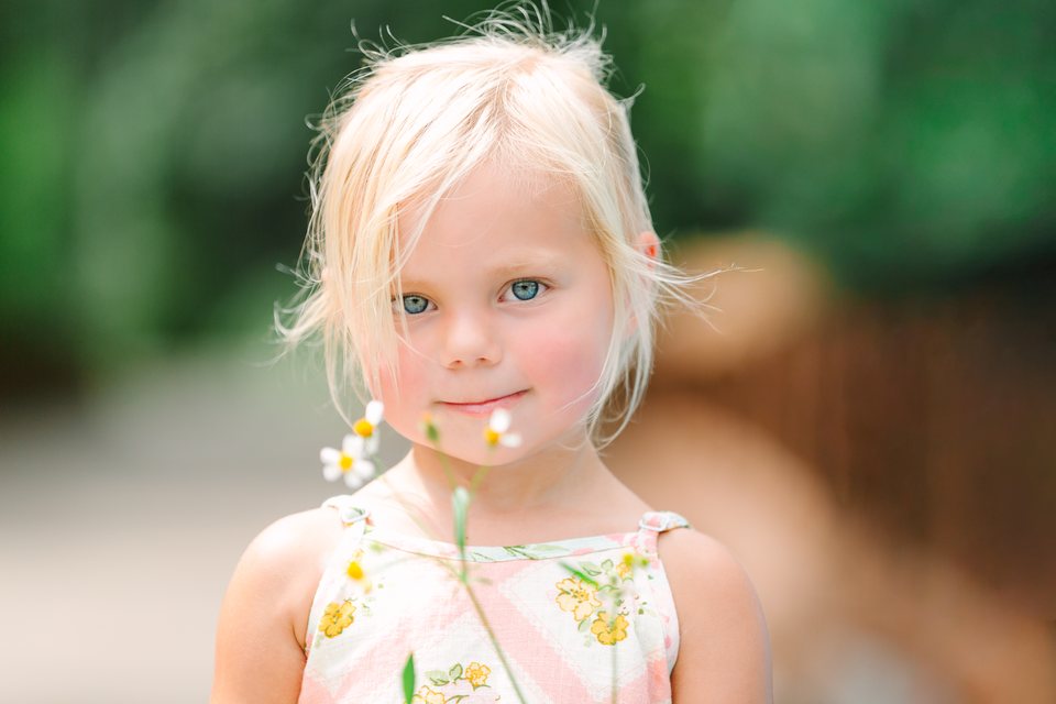 Toddler in a floral sundress holding a wildflower on a leafy path