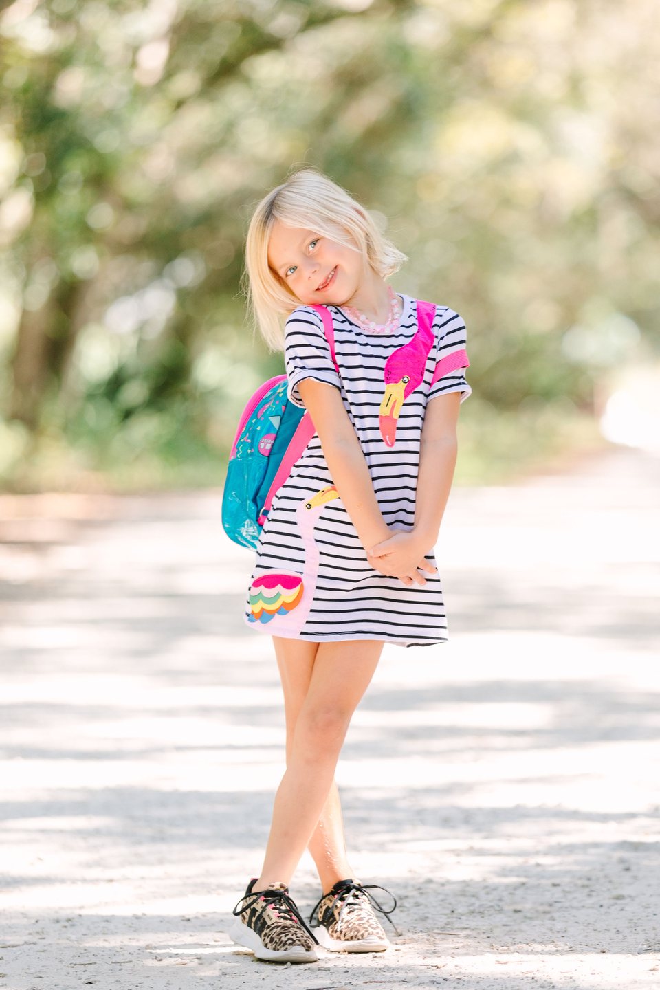 Smiling girl in a striped dress and backpack on a sunlit, tree-lined path