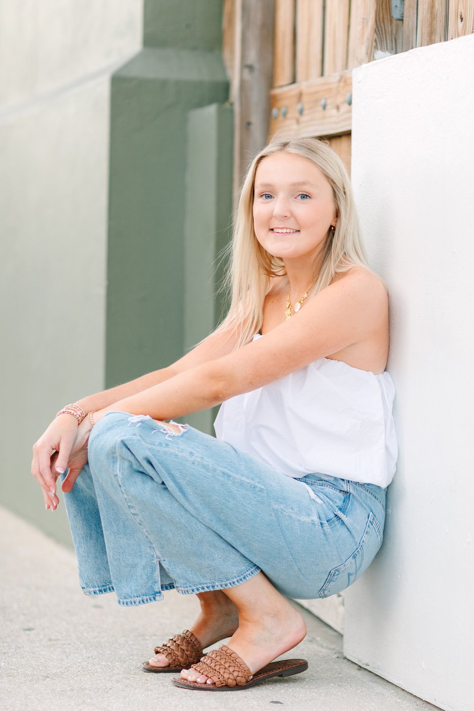 Young woman in a white top and jeans sitting against a white wall