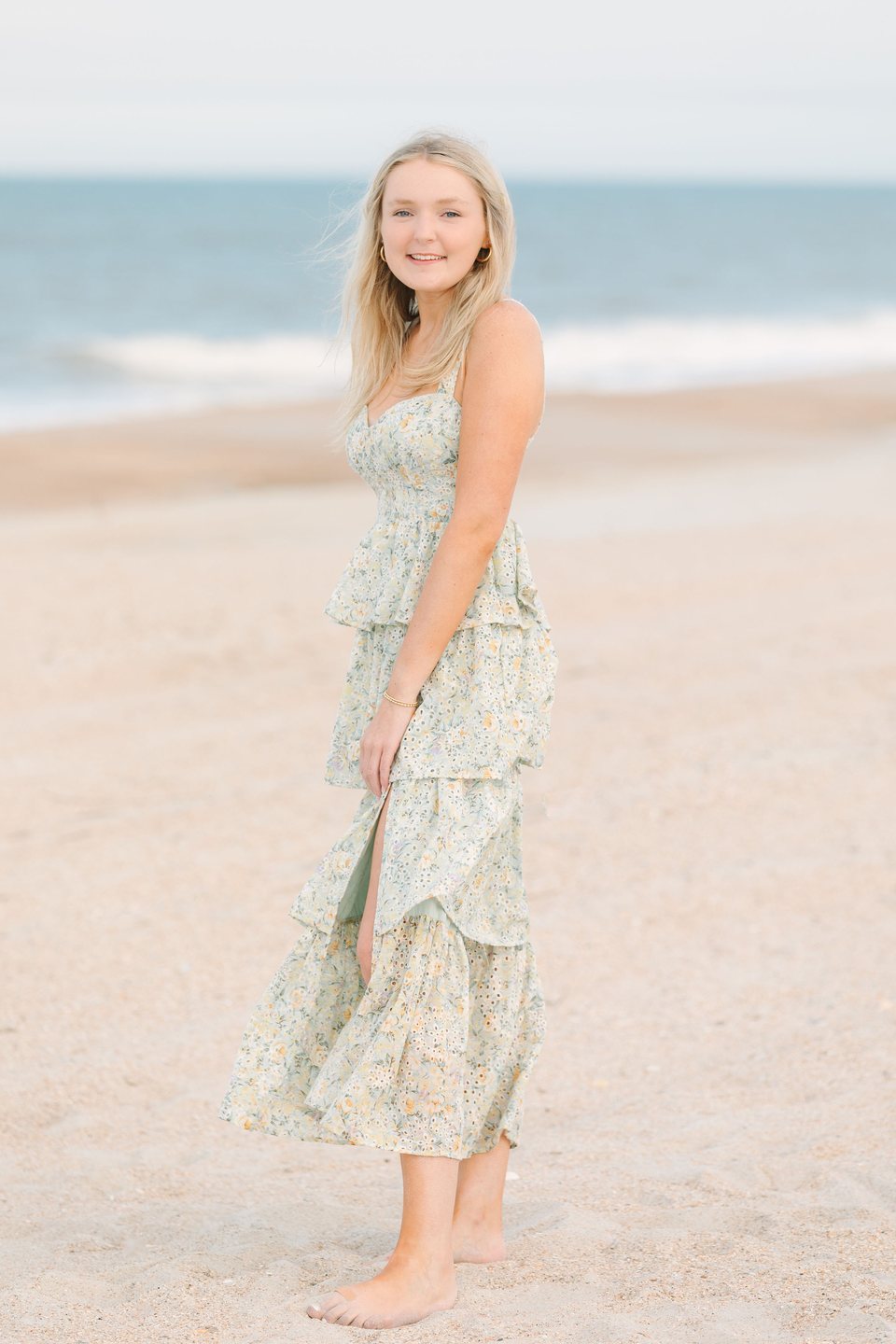 Young woman in a tiered floral dress standing barefoot on the beach
