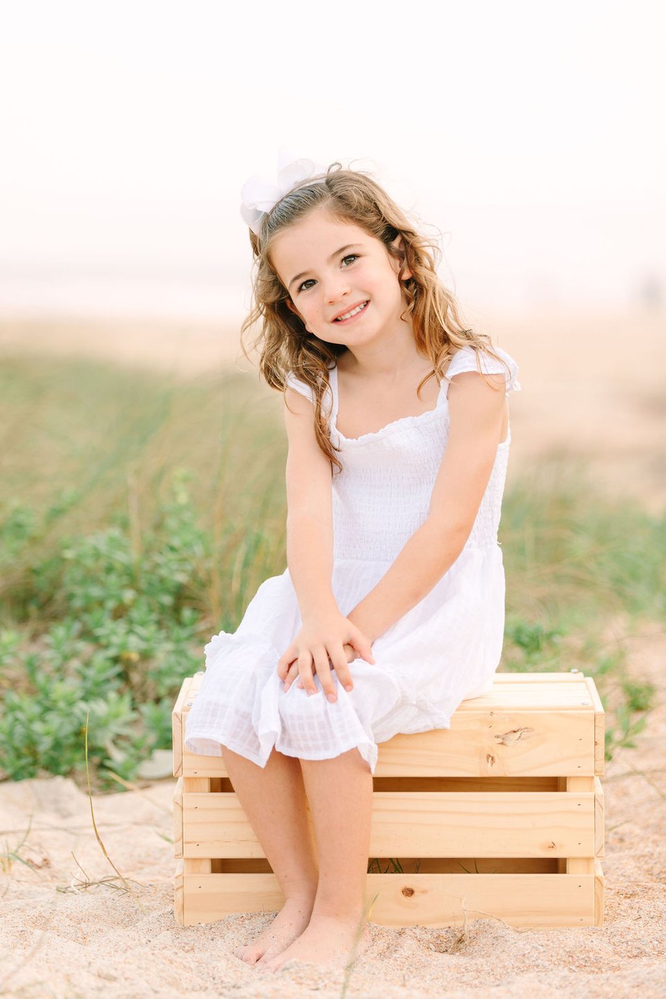 Child in a white dress sitting on a wooden crate at the beach
