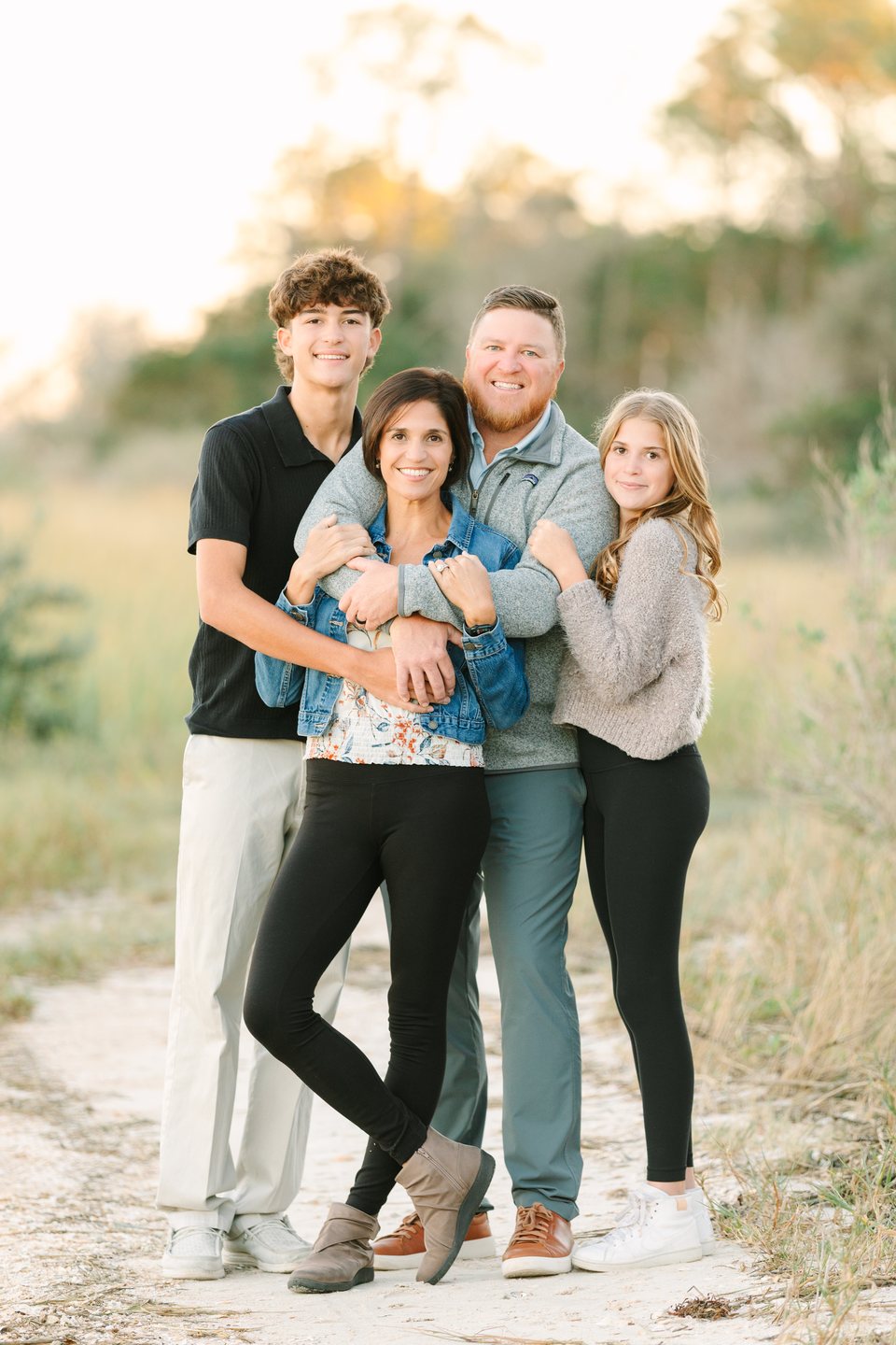 Family of four hugging and smiling on a sandy path among trees
