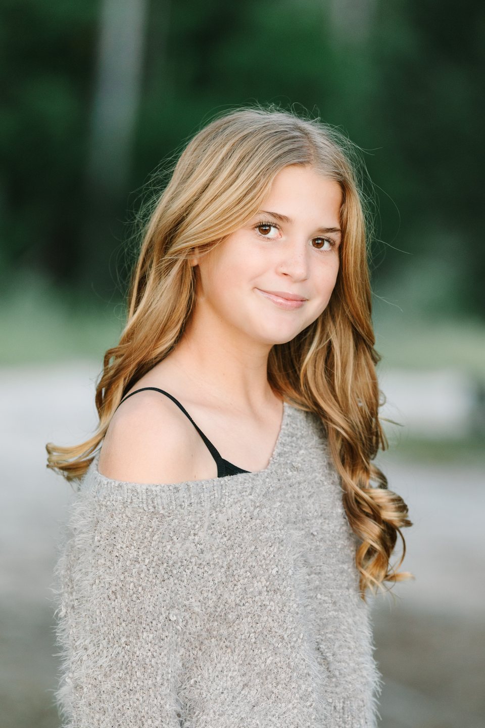 Teenage girl with long wavy hair in a gray sweater, smiling among green trees