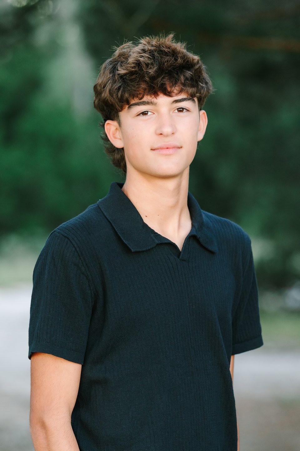 Close portrait of a teenage boy in a navy polo among dark green trees