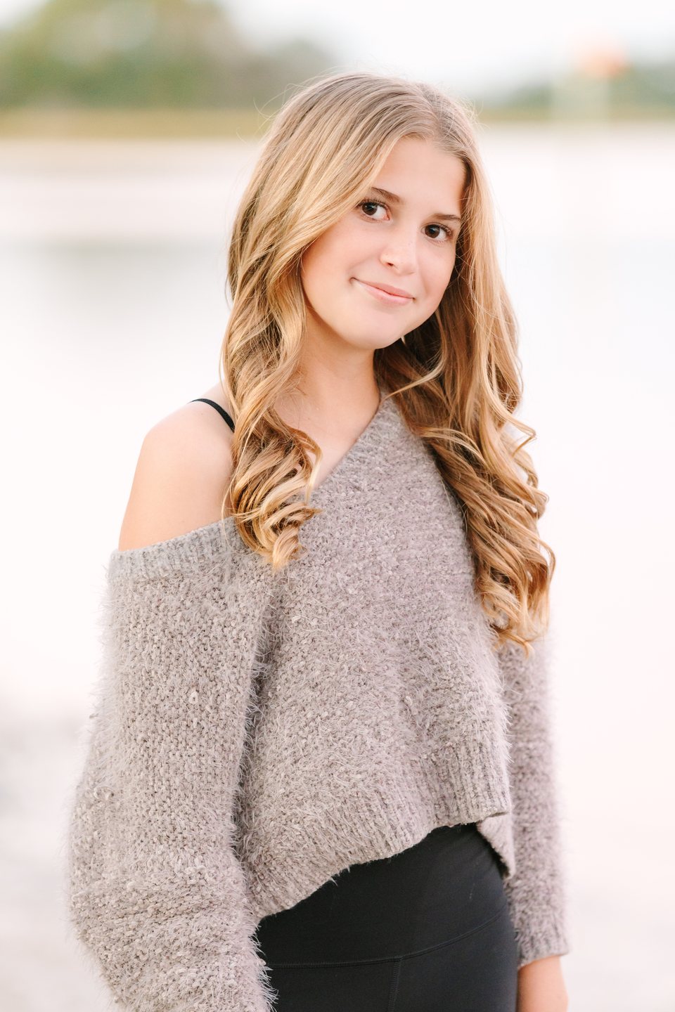 Teen in a gray sweater smiling beside the water at sunset