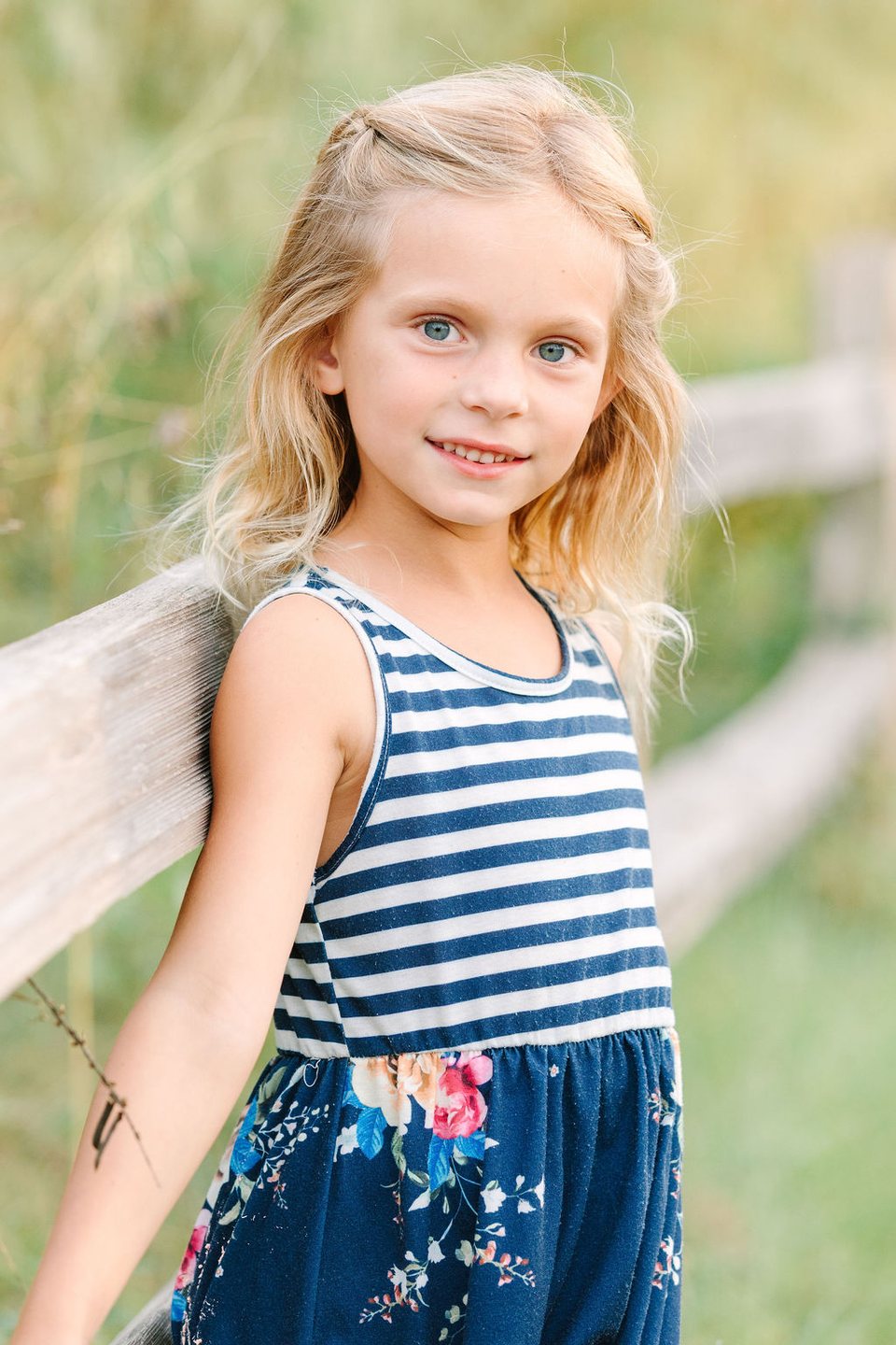 Girl in a striped top leaning on a wooden fence beside tall grass