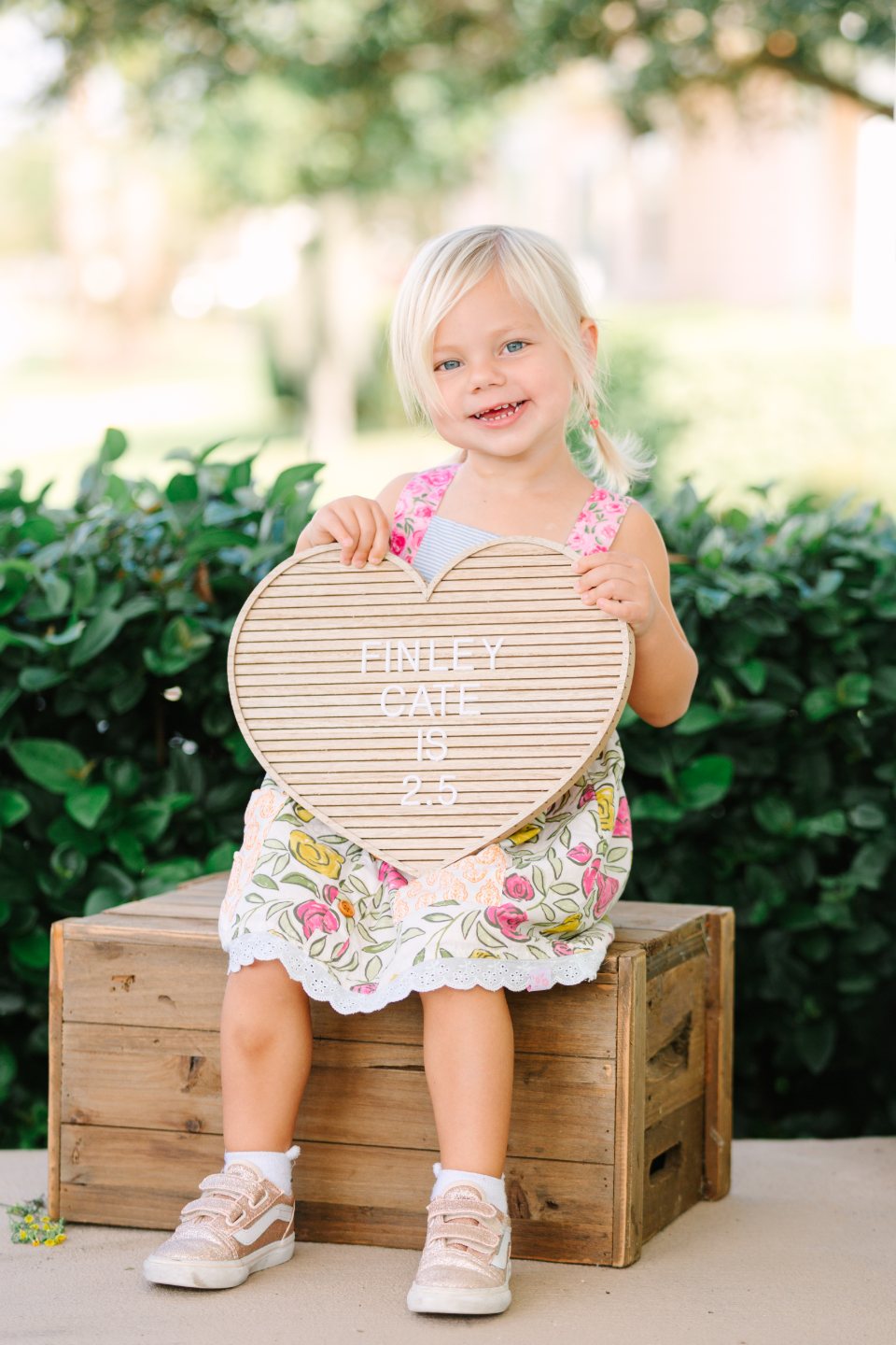 Smiling little girl on a wooden crate holding a heart-shaped milestone sign