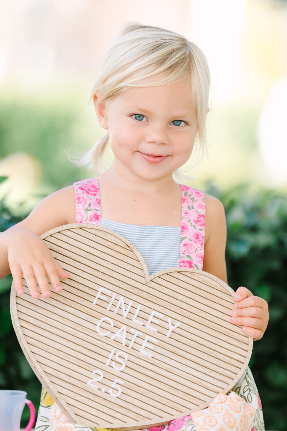 Smiling little girl holding a heart-shaped milestone sign among flowers