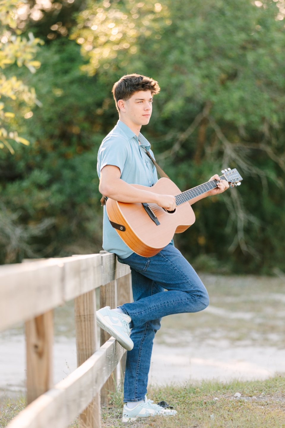 Teenage boy playing guitar while leaning on a wooden fence by the water