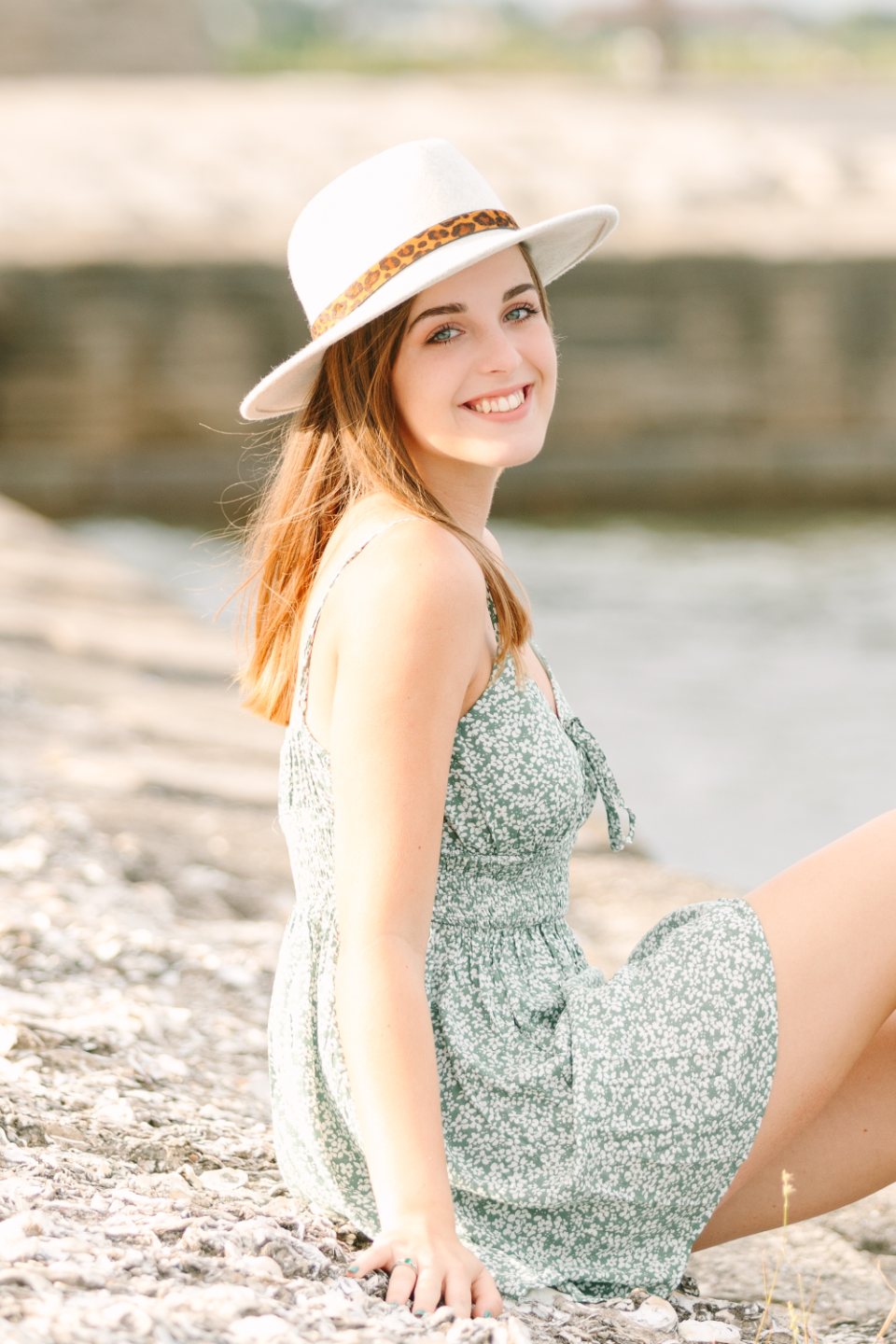 Young woman in a white hat and green sundress sitting on rocks by the water