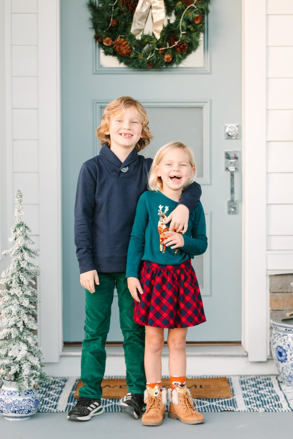 Two children in holiday outfits laughing by a front door with a wreath
