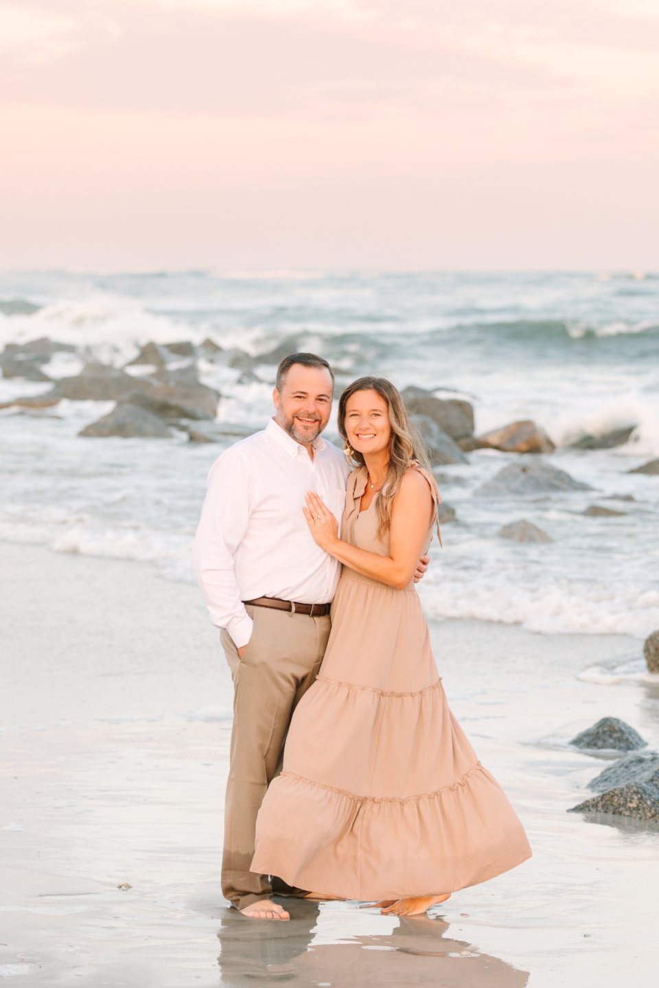 Couple embracing at the water's edge beside rocks at sunset