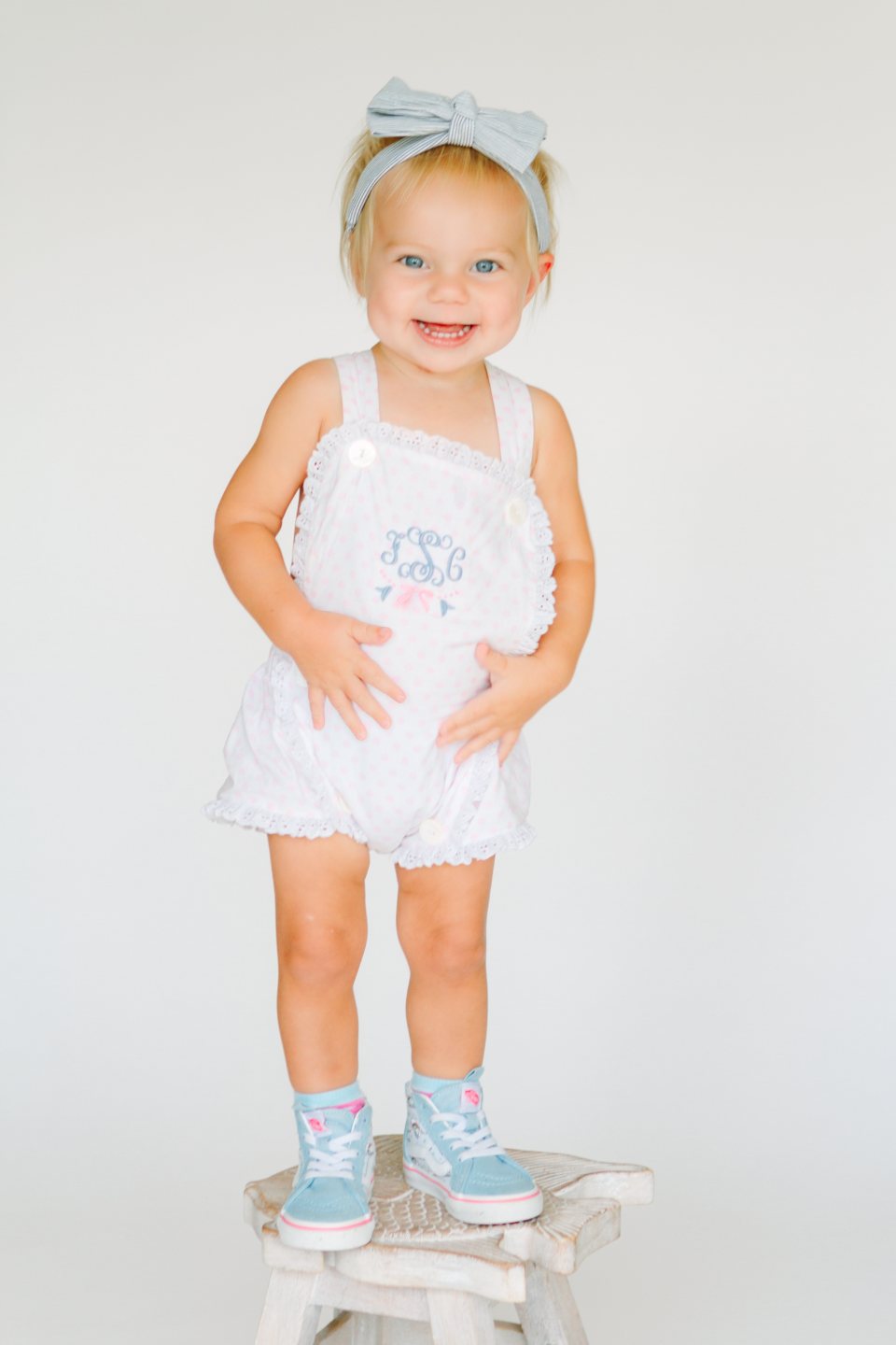 Toddler in a white romper and hair bow standing on a small stool in the studio