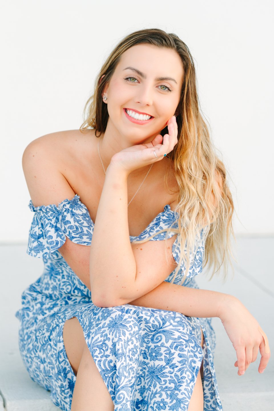 Young woman in a blue floral dress smiling with her chin on her hand in a white studio